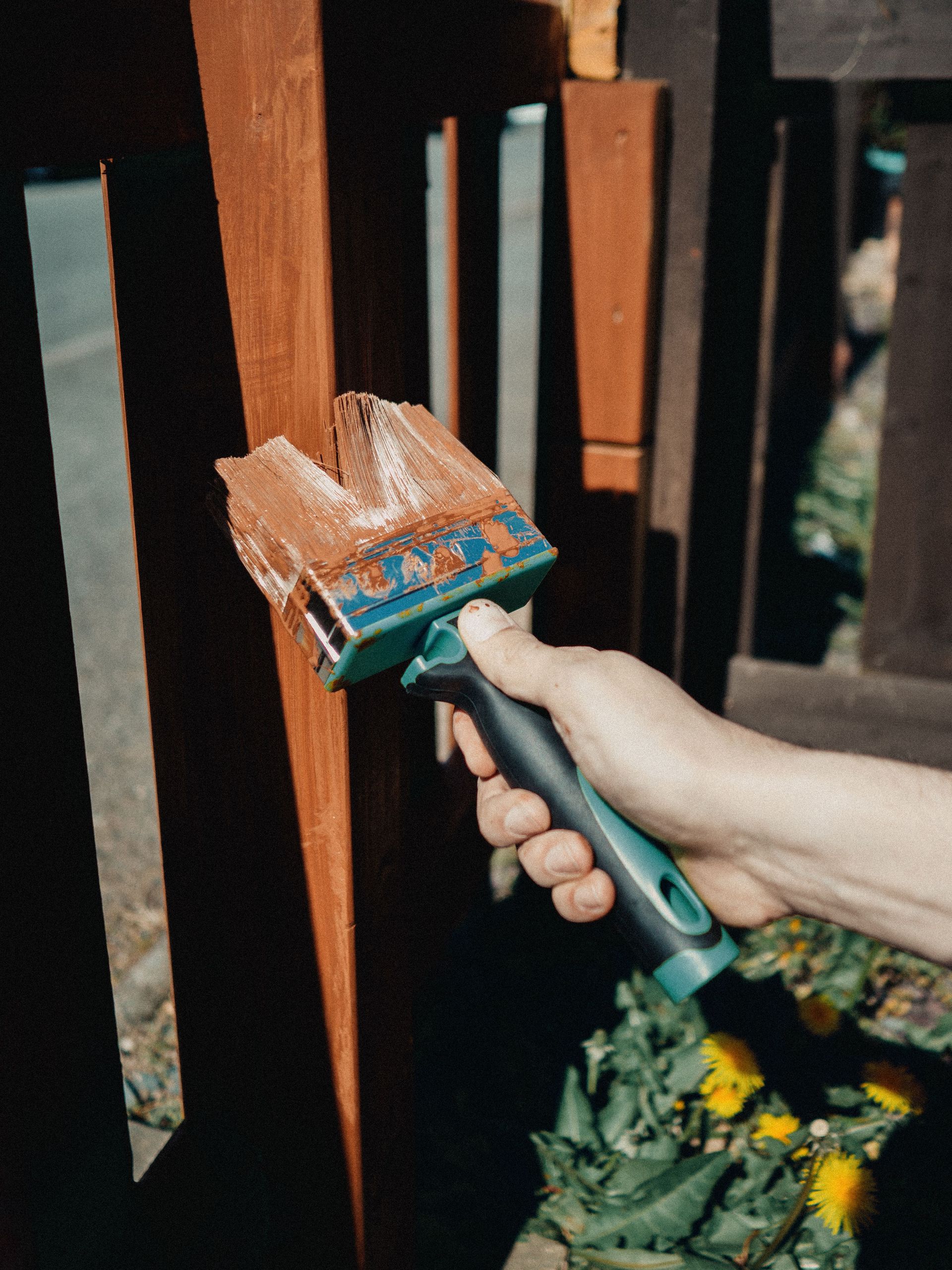 A close-up of a hand painting a wooden fence with a brush outdoors, surrounded by greenery.